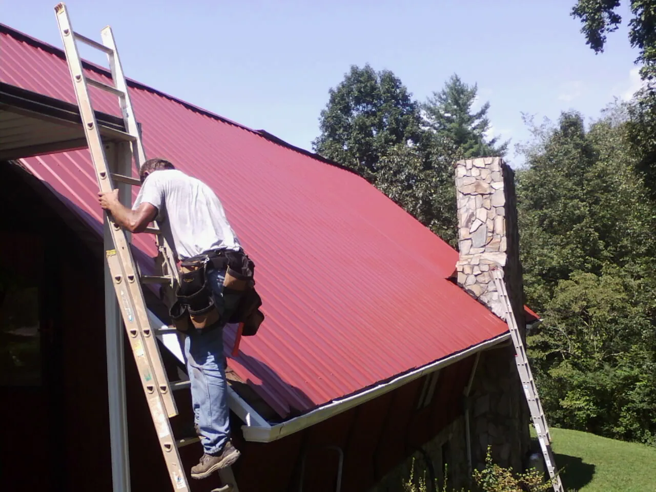 Roofer on a ladder beside a red metal roof and stone chimney — Soffit & Fascia Repair by Roofing Brothers in Jenkintown